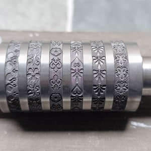 Könnte beinhalten: Eine Nahaufnahme einer Metallrolle mit fünf verschiedenen gravierten Mustern. Die Rolle ist silberfarben und hat eine glatte, polierte Oberfläche. Die Muster sind kompliziert und detailliert und scheinen für den Einsatz in einem Druck- oder Prägeprozess konzipiert zu sein.