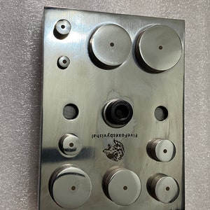 Può includere: Uno strumento per la formatura del metallo in acciaio inossidabile con varie rientranze circolari e un'apertura cilindrica centrale. Lo strumento ha una finitura spazzolata e il testo "FiveFoxesbyvishal" con un logo a testa di volpe.