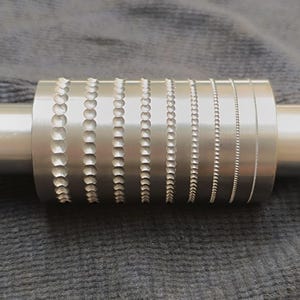 Könnte beinhalten: Eine silberne Metallwalze mit verschiedenen strukturierten Mustern, die in ihre Oberfläche geätzt sind. Die Muster umfassen Reihen von Kreisen, Kettengliedern und gepunkteten Linien. Die Walze ist zylindrisch und hat an jedem Ende einen glatten Griff.