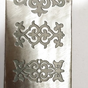 May include: A metal plate with four intricate, symmetrical designs cut out of it. The designs are in a traditional style, with swirls and curves.