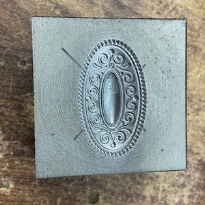 May include: A metal stamp with an oval design featuring intricate scrollwork and a beaded border.