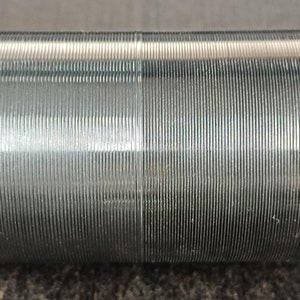 Könnte beinhalten: Zylindrische Metallkomponente mit geriffelter Oberfläche, montiert auf einer silbernen Stange. Die parallelen Rillen erzeugen ein strukturiertes Aussehen. Das Objekt ist wahrscheinlich Teil einer Maschine oder eines Mechanismus.