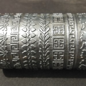 以下が含まれることがあります： 6つの異なるエンボス加工のデザインが施された銀色の金属ローラー。デザインには、渦巻き、花、ギリシャの鍵、ハート、葉、つるが含まれています。