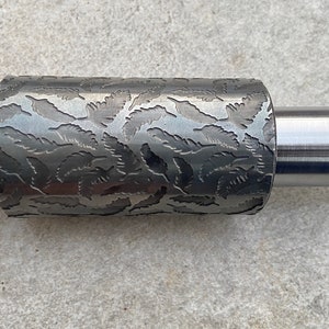 Könnte beinhalten: Ein silberner Nudelholz aus Metall mit einer strukturierten Oberfläche, die ein sich wiederholendes Muster aus stilisierten Blättern aufweist. Das Nudelholz hat an beiden Enden glatte, zylindrische Griffe.