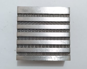 Może przedstawiać: Srebrny blok metalowy z sześcioma rzędami różnych wzorów w reliefie. Każdy rząd zawiera unikalny wzór, w tym kształty geometryczne, gwiazdy i inne elementy dekoracyjne.