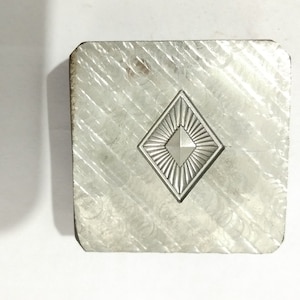Puede incluir: Un cuadrado de metal plateado con un diseño en forma de diamante en el centro. El diamante tiene una superficie texturizada en relieve y un diamante más pequeño en el centro.