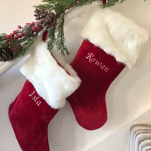 May include: Two deep red velvet Christmas stockings featuring white faux fur cuffs. The left stocking is embroidered with the name Isla in white script, and the right stocking is embroidered with the name Rowan in white script, hanging from a garland.