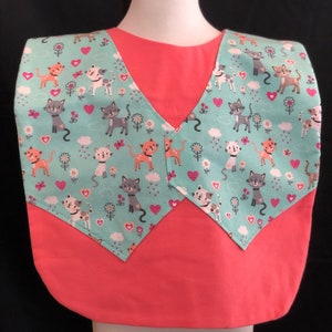 May include: A pink sleeveless shirt with a turquoise bib featuring a pattern of cats, hearts, and flowers. The bib has a V-neckline and is attached to the shirt with a seam.