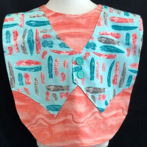 May include: A turquoise and coral colored shirt with a feather print. The shirt has a V-neck and two green buttons.