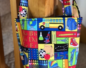 May include: A colorful tote bag with a school theme. The bag features a school bus, crayons, a globe, a laptop, a soccer ball, and the letters of the alphabet. The bag is made of fabric and has a blue lining.
