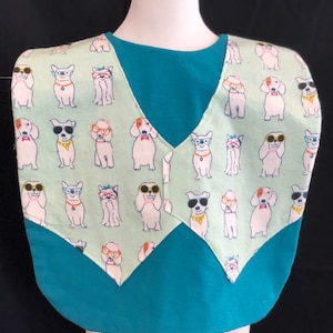 May include: A turquoise bib with a green pattern of dogs wearing sunglasses. The bib has a V-neck and a pointed bottom.