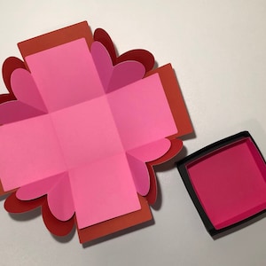 May include: A pink and red paper box with a heart-shaped design. The box is open and the inside is visible. The box is made of paper and has a black base.