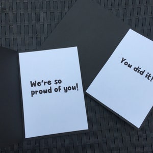 May include: A white card with black text that reads "We're so proud of you!" and a second white card with black text that reads "You did it!" Both cards are on a black surface.