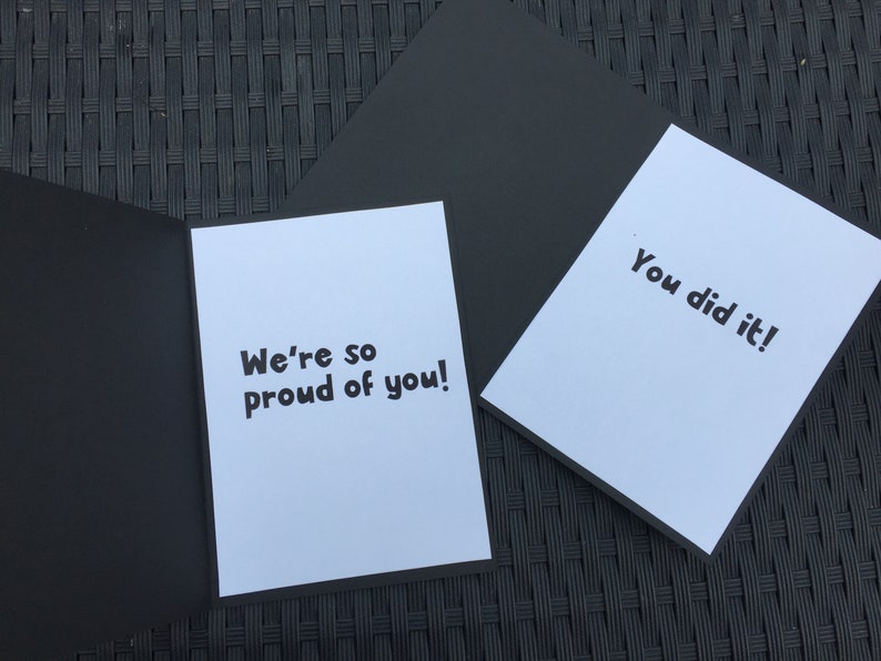 May include: A white card with black text that reads "We're so proud of you!" and a second white card with black text that reads "You did it!" Both cards are on a black surface.