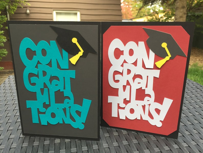 May include: Two graduation cards with the words "Congratulations" in different colors and styles. The cards are black with a red and a teal background. Each card has a black graduation cap with a yellow tassel.