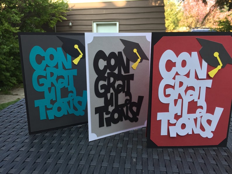May include: Three graduation cards with the words "Congratulations" cut out in different colors. The cards are on a black wicker surface.