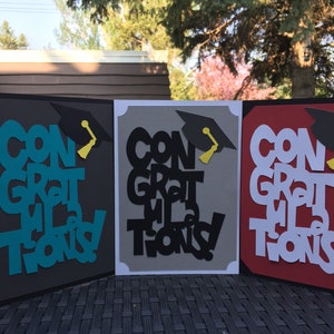 May include: Three graduation cards with the words "Congratulations" in different colors and styles. The cards are decorated with a black graduation cap and a yellow tassel.