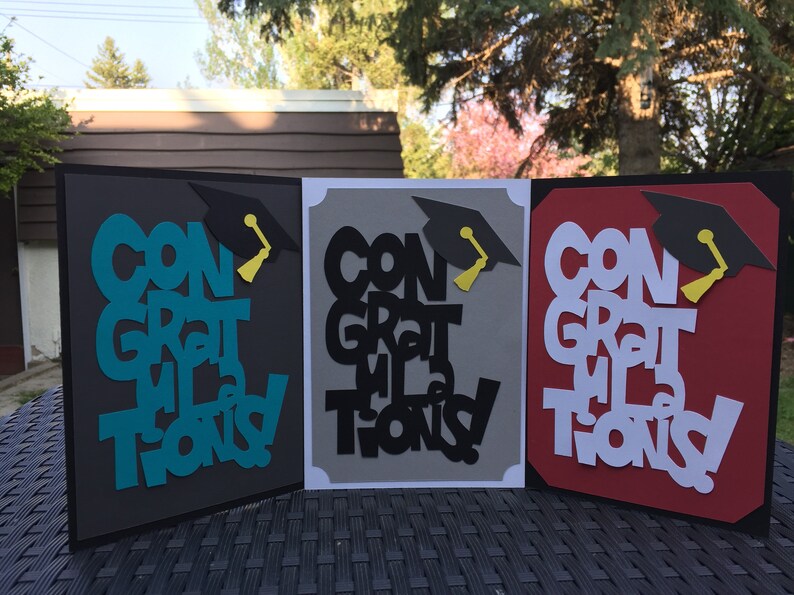 May include: Three graduation cards with the words "Congratulations" in different colors and styles. The cards are decorated with a black graduation cap and a yellow tassel.
