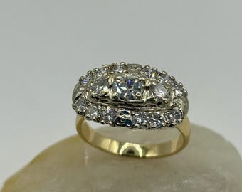 May include: A gold ring with a large, clear center stone surrounded by smaller clear stones. The ring is set on a white stone.
