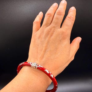 May include: A red bangle bracelet with silver-colored floral designs. The bracelet is worn on a wrist, showcasing its glossy finish and decorative elements. The bracelet is a piece of jewelry.