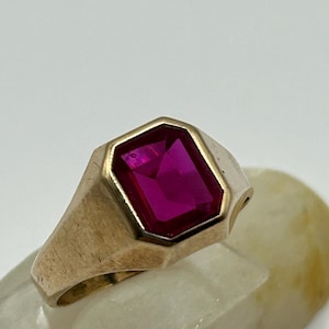 以下が含まれることがあります： 大きな長方形の濃いピンク色の宝石をあしらったゴールドトーンのリング。リングは、太いバンドと宝石用のベゼルセッティングを備えた、クラシックなシグネットスタイルのデザインです。宝石はファセット加工されており、光を捉えて鮮やかな輝きを生み出しています。