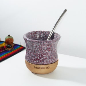 Può includere: Una tazza da mate in ceramica con smalto maculato bordeaux e azzurro, dotata di una base in legno naturale incisa con la parola MATECITO. Una sottile cannuccia bombilla in acciaio inossidabile lucido riposa nella tazza dal bordo ampio e svasato.