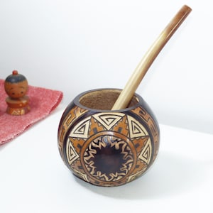 Può includere: Una tazza da mate rotonda in legno con un design geometrico nei toni del marrone e beige, con una cannuccia di legno. Due piccole bambole di legno e un panno rosso sono sullo sfondo. La tazza ha un diametro di circa 10 cm.