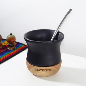 Può includere: Un contenitore per mate nero opaco con una base in legno chiaro naturale che riporta il testo inciso MATECITO. Una cannuccia bombilla in metallo argentato è inserita nel contenitore. Il recipiente ha una forma curva a bordo largo su fondo bianco.