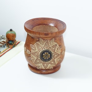 Può includere: Un contenitore per mate in legno marrone con un'ampia apertura arrotondata e una base affusolata. L'esterno è decorato con un motivo geometrico circolare intagliato che include motivi a forma di foglia e un fiore centrale a quattro petali.
