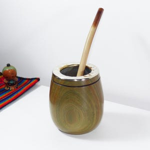 Può includere: Un contenitore per mate in legno arrotondato con un bordo in metallo color argento lucido sull'apertura superiore. Una cannuccia in legno lunga, sottile e di colore chiaro con una punta marrone scuro è inserita al centro del contenitore in legno.