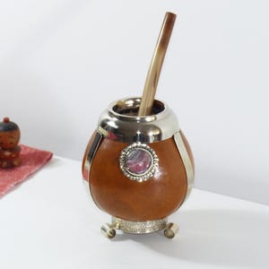 Può includere: Una zucca di cuoio marrone con accenti argentati e un'intarsio di pietra decorativa. La zucca è su un supporto con tre gambe e ha una cannuccia di bombilla di legno che sporge dalla parte superiore.