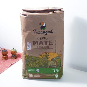 Peut inclure: Un sac en papier brun de Tucanguá Yerba Mate Organica, avec un logo de toucan. Le sac présente une illustration verte et marron de plantes et le texte "Yerba Mate Organica". Le sac pèse 1 kg.
