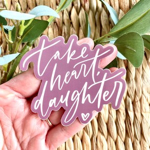 May include: A purple vinyl sticker with white lettering that reads "Take heart, daughter" with a small white heart at the bottom.