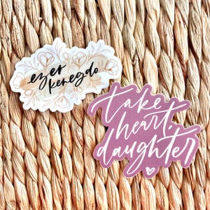 May include: Two stickers with inspirational quotes. The first sticker is white with a watercolor background and gold floral accents. The quote is "ever  kenegdo". The second sticker is purple with white lettering. The quote is "take heart daughter".