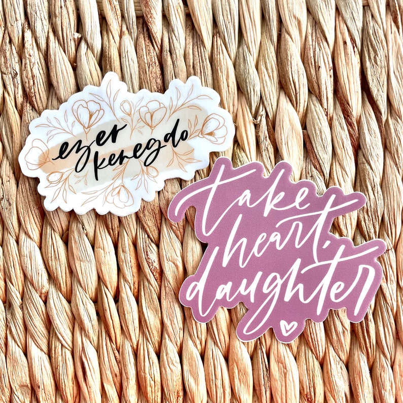 May include: Two stickers with inspirational quotes. The first sticker is white with a watercolor background and gold floral accents. The quote is "ever  kenegdo". The second sticker is purple with white lettering. The quote is "take heart daughter".