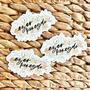 K&ouml;nnte beinhalten: Drei wei&szlig;e Aufkleber mit floralen Designs und dem Text "ezer keregdo" in schwarzer Schrift. Die floralen Elemente sind hellbraun. Die Aufkleber befinden sich auf einer gewebten, hellbraunen Oberfl&auml;che.