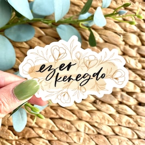 K&ouml;nnte beinhalten: Ein wei&szlig;er Aufkleber mit dem Schriftzug "ezer kenedo" in schwarzer Schrift, umgeben von einem goldenen Blumendesign. Der Aufkleber hat einen beigefarbenen Aquarell-Hintergrund und liegt auf einer gewebten Oberfl&auml;che.