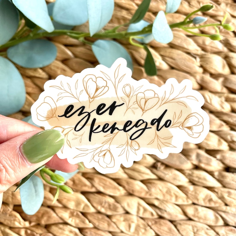K&ouml;nnte beinhalten: Ein wei&szlig;er Aufkleber mit dem Schriftzug "ezer kenedo" in schwarzer Schrift, umgeben von einem goldenen Blumendesign. Der Aufkleber hat einen beigefarbenen Aquarell-Hintergrund und liegt auf einer gewebten Oberfl&auml;che.