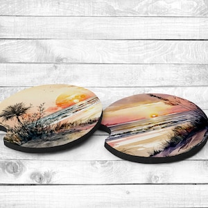 May include: Two round beverage coasters with a watercolor sunset scene over the ocean. The coasters feature a palm tree, sand, and waves.