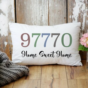 May include: White throw pillow with the numbers 97770 in different colors and the text "Home Sweet Home" in black cursive font. The pillow is on a wooden floor with a gray knitted blanket and a vase of pink flowers.