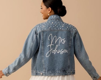 Può includere: Giacca di jeans blu chiaro con perle sulla parte superiore della schiena. La giacca ha il nome "Mrs Johnson" in corsivo bianco sul retro. La giacca è indossata sopra un vestito bianco.