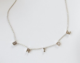 May include: A delicate silver necklace spelling out "BRIDE" in individual letter charms. The necklace is displayed on a white surface, alongside a beaded clutch and a decorative dish.