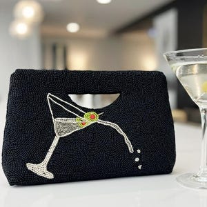 K&ouml;nnte beinhalten: Schwarze, perlenbesetzte Clutch mit Martini-Glas-Design. Die Clutch hat einen Griff und ein Martini-Glas mit Oliven. Ein Martini-Glas mit Oliven befindet sich neben der Clutch.