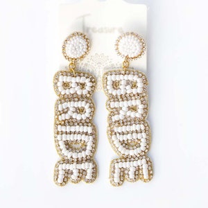 May include: A pair of gold beaded earrings with the word "BRIDE" spelled out in white beads.