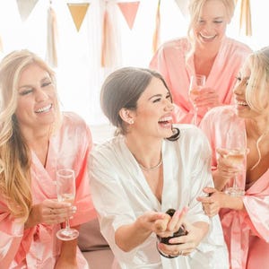 Può includere: Quattro donne in vestaglie di raso rosa e bianco stanno festeggiando con lo champagne. Sorridono e ridono insieme. La donna in vestaglia bianca sta aprendo una bottiglia di champagne.