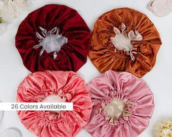May include: Four circular satin hair bonnets in deep red, copper, coral, and light pink, each featuring a gathered elastic edge and a contrasting light-colored interior lining. The text 26 Colors Available is printed on a white rectangular overlay.