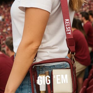 A person wearing a white t-shirt and blue jeans is holding a red purse with a clear plastic window, which has the text "GIG 'EM" printed on it.
