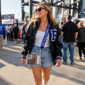 May include: A clear rectangular stadium bag with a blue strap featuring the white text GO FALCONS! GO. The bag contains a smartphone, a brown checkered wallet, an orange lipstick, and a white ticket. It is worn with a black varsity jacket.