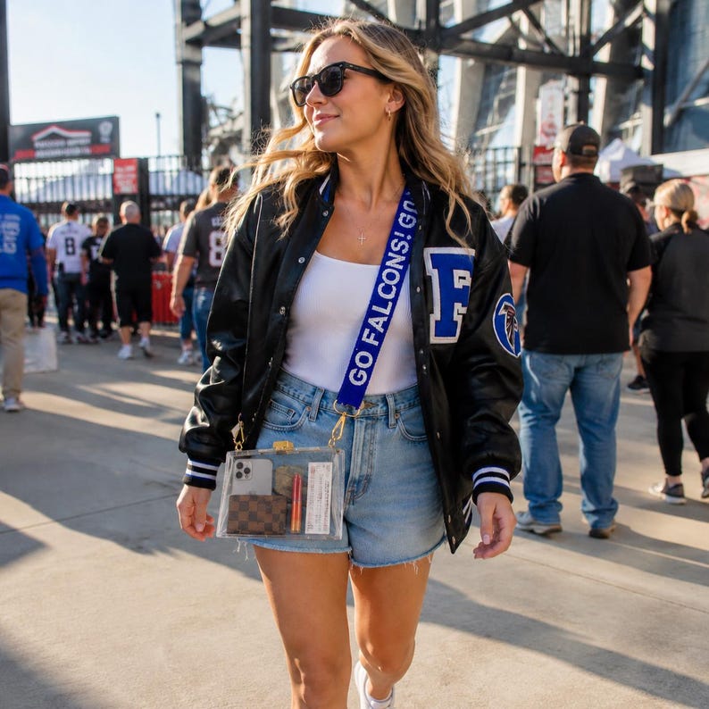 May include: A clear rectangular stadium bag with a blue strap featuring the white text GO FALCONS! GO. The bag contains a smartphone, a brown checkered wallet, an orange lipstick, and a white ticket. It is worn with a black varsity jacket.