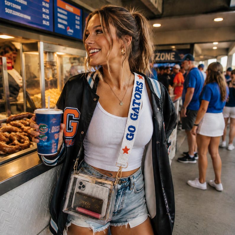 May include: A clear plastic crossbody bag with a white strap featuring the blue text GO GATORS and a small orange star. The bag contains a smartphone, a brown patterned wallet, and a pink lipstick, worn over a white ribbed tank top and denim shorts.
